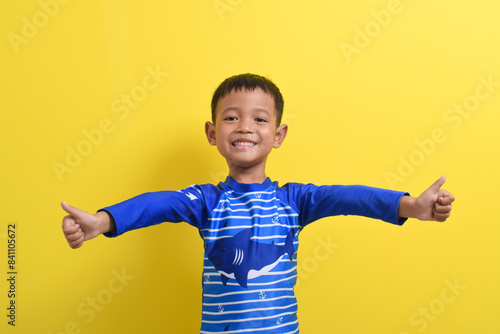 Portrait of a cute little Asian boy wearing a swimsuit shows thumbs up isolated on a yellow background. Summer vacation concept. With Copy space