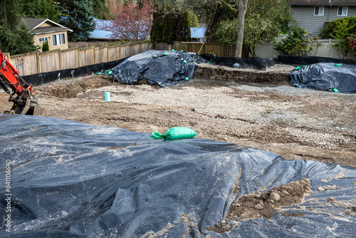 Beginnings of a new house, foundation excavation started with backhoe heavy equipment, hole in the ground with black tarps covering piles of dirt
