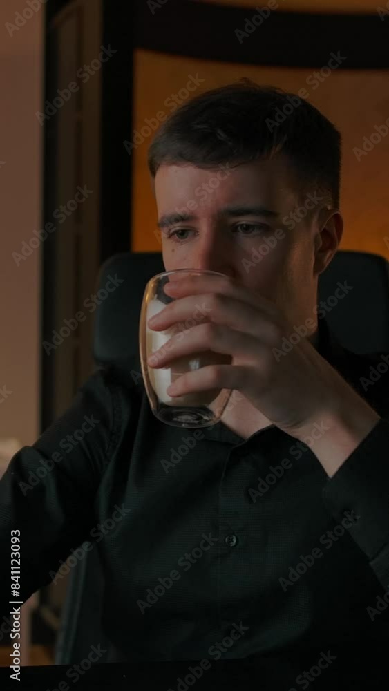 A young caucasian man drinks coffee at home in the evening at his laptop. Beautiful cozy interior, a man drinks coffee from a transparent glass cup, passionate about content from the Internet A young caucasian man drinks coffee at home in the evening at his laptop. Beautiful cozy interior, a man drinks coffee from a transparent glass cup, passionate about content from the Internet