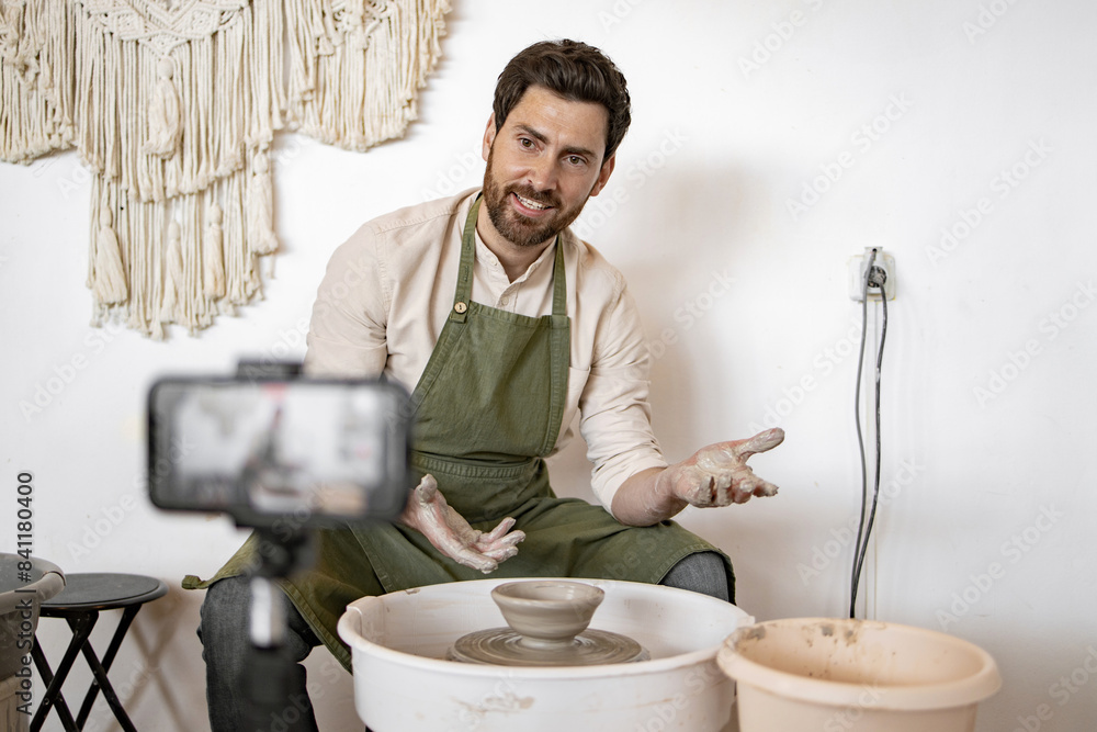 Male potter sculpting a ceramic vase using a potter's wheel while recording a video blog on smartphone.