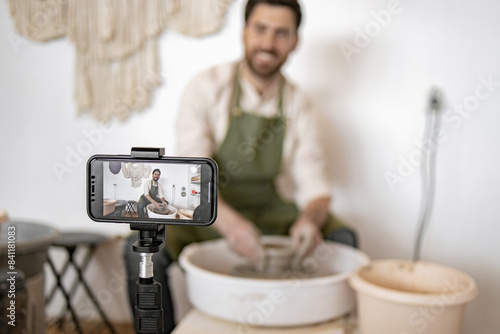 Adult male potter creating pottery on a potter's wheel while recording a video blog using a smartphone. Focus on ceramic art and social media content creation.