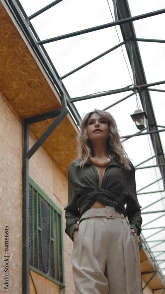 Low angle of positive woman in stylish elegant outfit holding hands in pockets and looking away while posing for camera in corridor of barn. Vertical shot. Low angle of positive woman in stylish elegant outfit holding hands in pockets and looking away while posing for camera in corridor of barn. Vertical shot.