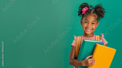 Obraz na płótnie A cheerful young school girl with a backpack, smiling and holding colorful books against a green background. Perfect image for back-to-school themes. w salonie