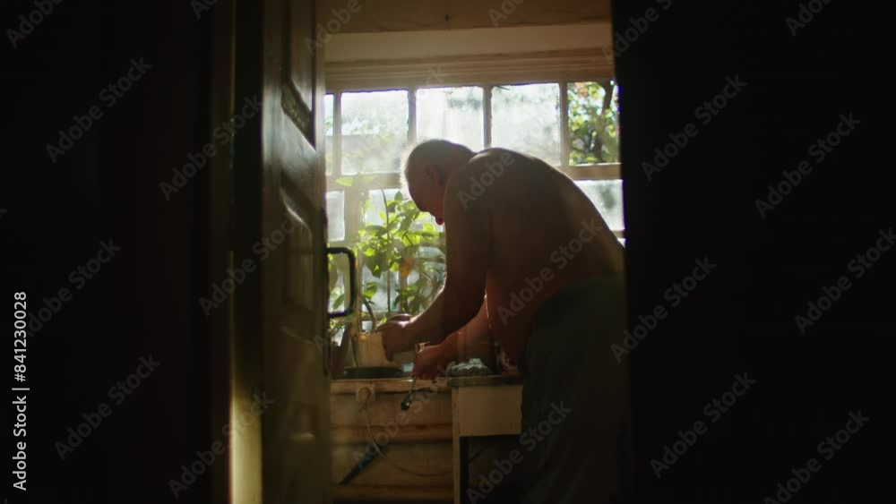 Older man standing by a window preparing food. Natural light from windows in the background. Domestic life and morning routine concept. Design for poster, banner, greeting card. Candid shot. Older man standing by a window preparing food. Natural light from windows in the background. Domestic life and morning routine concept. Design for poster, banner, greeting card. Candid shot.