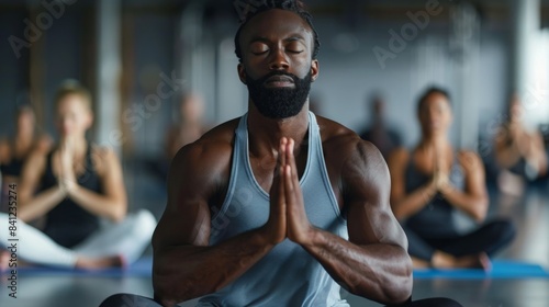 A man meditates with his eyes closed and hands in prayer position, participating in a yoga class with other people in the background.