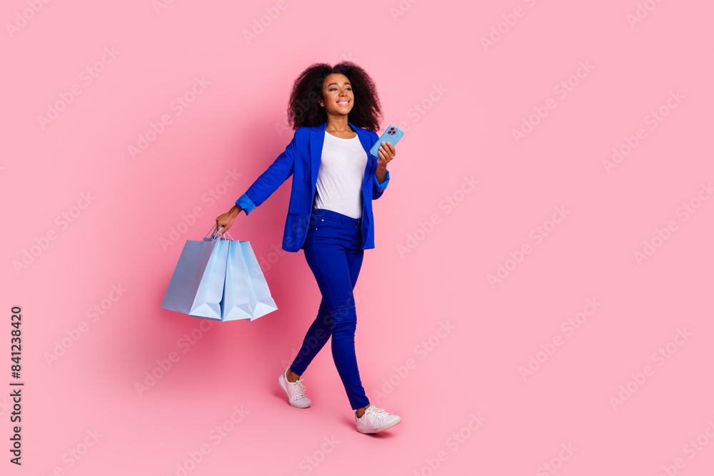 Full length photo of positive woman wear blue jacket hold shopping bags smartphone look empty space isolated on pink color background Full length photo of positive woman wear blue jacket hold shopping bags smartphone look empty space isolated on pink color background