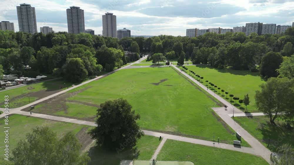 Park during a beautiful summer day surrounded by lush greenery, grass, and trees under a blue sky with buildings in the distance Park during a beautiful summer day surrounded by lush greenery, grass, and trees under a blue sky with buildings in the distance