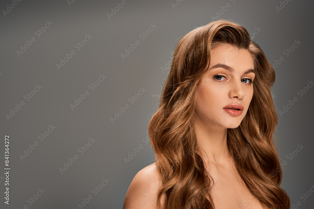 Naklejka premium A young beautiful woman with long, wavy hair elegantly posing on gray backdrop
