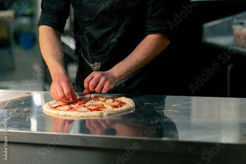 in a professional kitchen in a pizzeria young chef in a black uniform puts ingredients on pizza