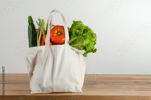 Fresh vegetables like lettuce, zucchini, carrots and tomatoes are coming out of a white reusable bag placed on a wooden table with a white background photo