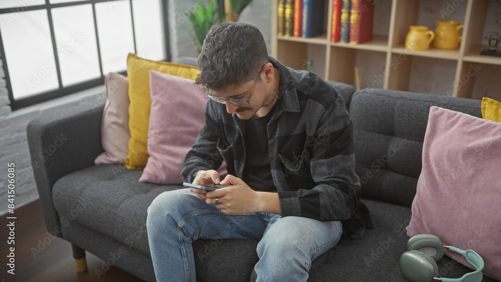 A young hispanic man using a smartphone on the couch in a cozy living room. A young hispanic man using a smartphone on the couch in a cozy living room.