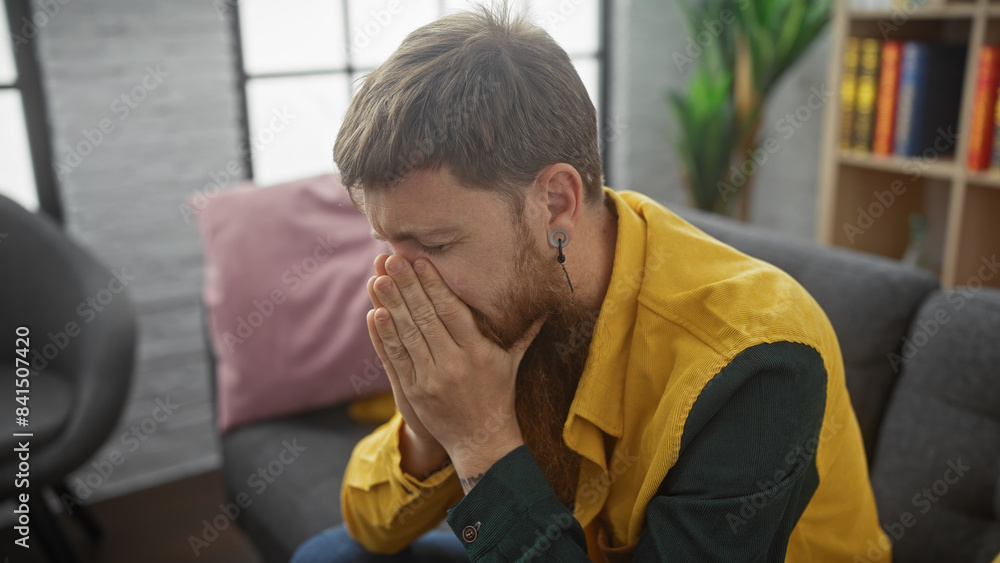 A distraught bearded redhead man sitting indoors, covering face with hands in apparent distress or sadness. A distraught bearded redhead man sitting indoors, covering face with hands in apparent distress or sadness.