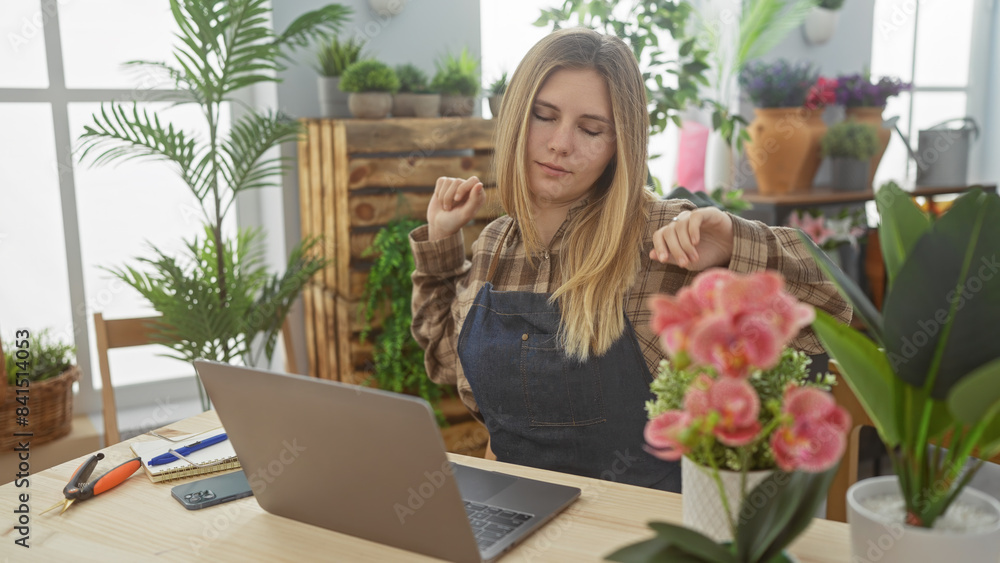 A young woman florist with blonde hair stretches during a break in a green, plant-filled indoor flower shop. A young woman florist with blonde hair stretches during a break in a green, plant-filled indoor flower shop.