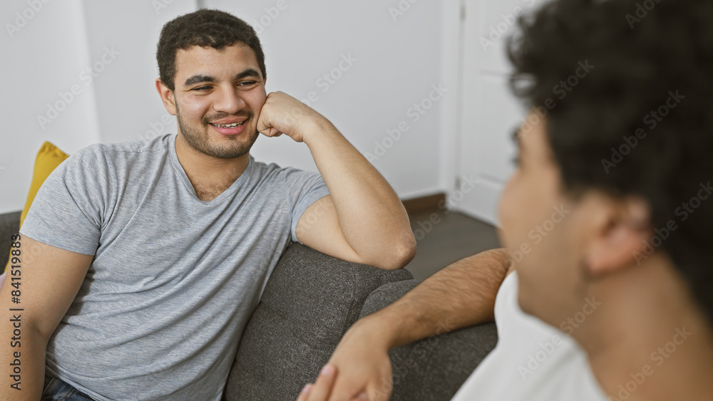 Two men relax on the couch, engaging in a friendly conversation, embodying warmth and connectivity in a casual home setting. Two men relax on the couch, engaging in a friendly conversation, embodying warmth and connectivity in a casual home setting.
