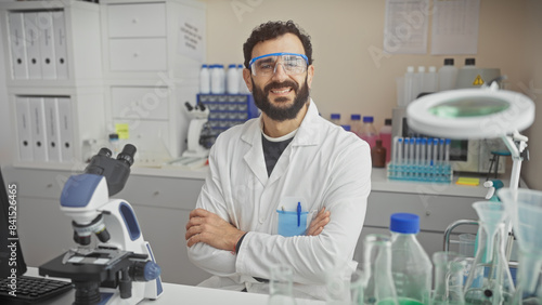 Billede på lærred Smiling bearded man in lab coat, safety goggles, standing in lab with microscope and scientific equipment