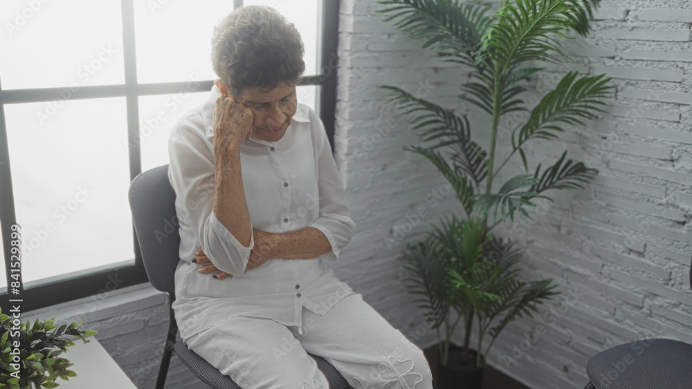 Elderly woman with short hair sits pensively in a white shirt and pants in an indoor waiting room with plants and a large window. Elderly woman with short hair sits pensively in a white shirt and pants in an indoor waiting room with plants and a large window.