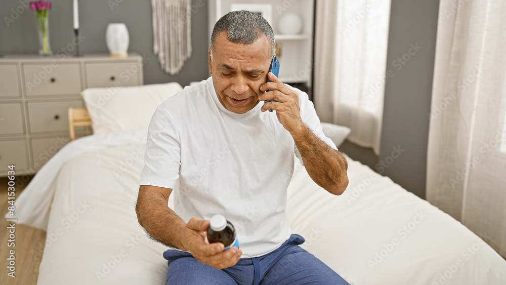 A middle-aged hispanic man in casual attire talking on a phone while holding a medicine bottle, seated on a bed in a well-lit bedroom. A middle-aged hispanic man in casual attire talking on a phone while holding a medicine bottle, seated on a bed in a well-lit bedroom.
