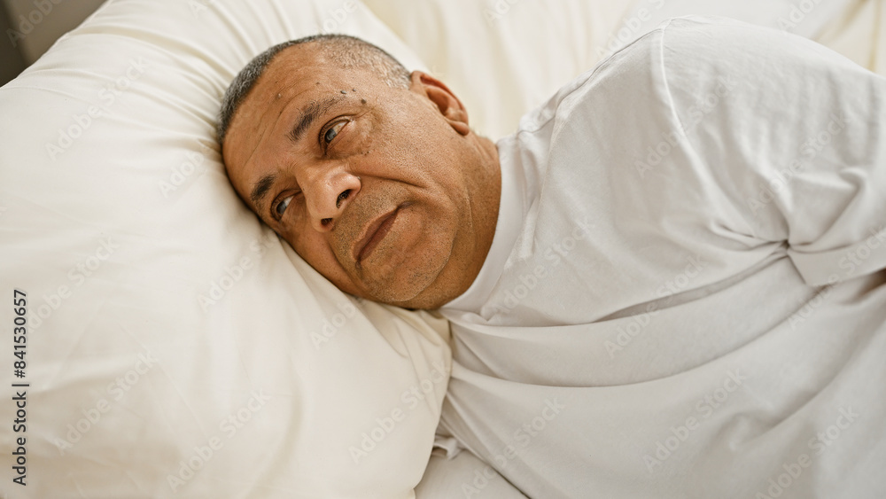 A middle-aged hispanic man appears contemplative while resting in a bedroom with white bedding. A middle-aged hispanic man appears contemplative while resting in a bedroom with white bedding.