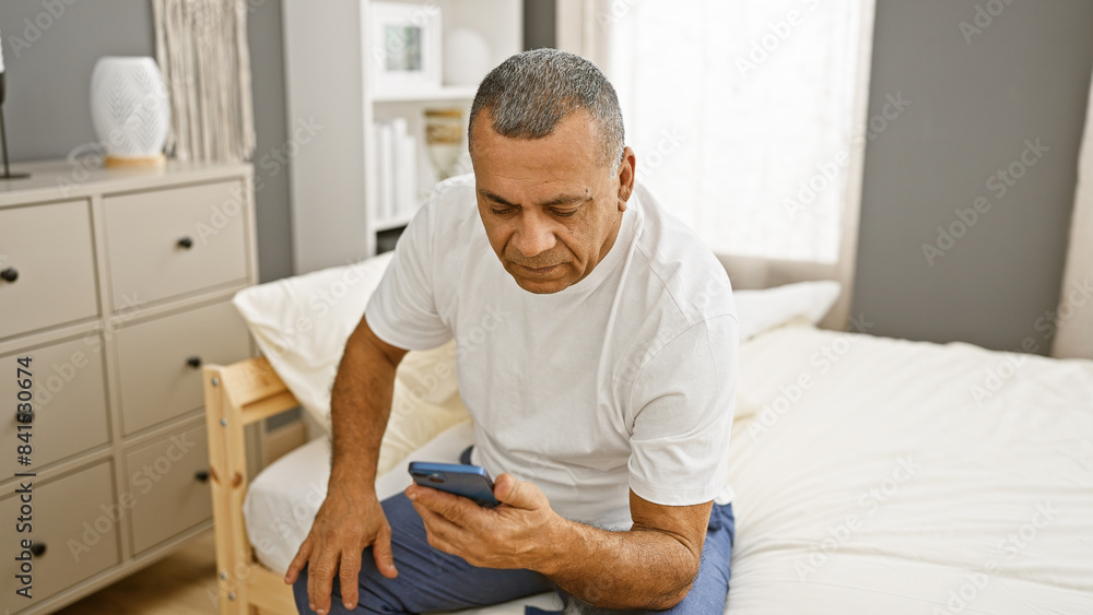 Mature hispanic man in white shirt sitting on bed with smartphone in bedroom interior. Mature hispanic man in white shirt sitting on bed with smartphone in bedroom interior.