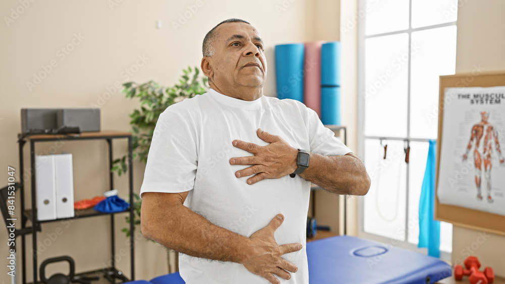 Obraz premium Middle-aged hispanic man experiencing discomfort in a well-equipped physical therapy clinic.