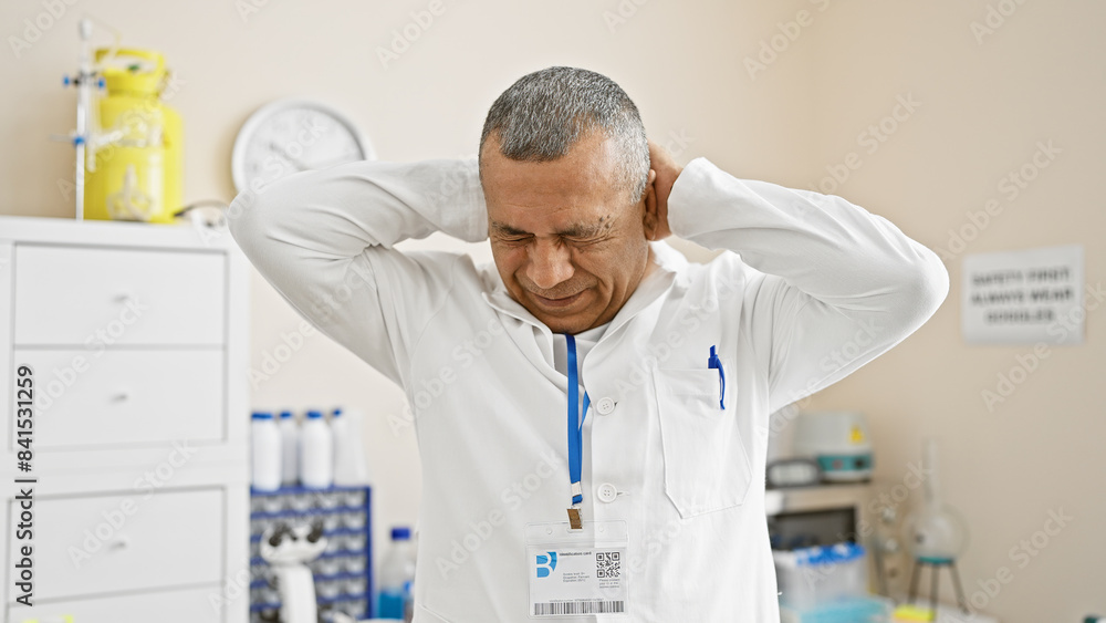 A middle-aged hispanic man in lab attire expresses stress or headache in a clinical hospital setting. A middle-aged hispanic man in lab attire expresses stress or headache in a clinical hospital setting.