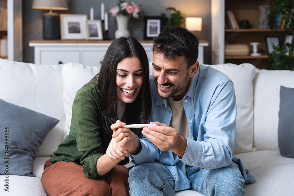 Expectations, wanted pregnancy. Young happy couple man and woman sitting at home on sofa holding and looking at pregnancy test. New life concept, waiting for positive pregnant result. Simple living. Expectations, wanted pregnancy. Young happy couple man and woman sitting at home on sofa holding and looking at pregnancy test. New life concept, waiting for positive pregnant result. Simple living.
