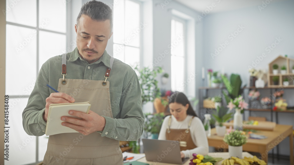 Man and woman working together in a bright flower shop, with the man writing and woman using a laptop. Man and woman working together in a bright flower shop, with the man writing and woman using a laptop.