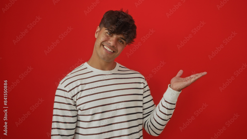 Fototapeta premium Handsome young hispanic man presents with an extended hand against an isolated red background.