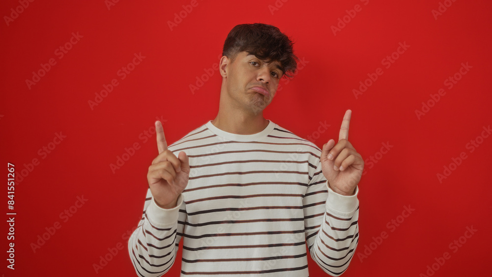 Portrait of a handsome young hispanic man in a striped shirt over an isolated red background, pointing upwards with both hands. Portrait of a handsome young hispanic man in a striped shirt over an isolated red background, pointing upwards with both hands.
