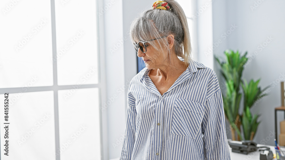 Mature woman with grey hair in a striped shirt stands thoughtfully in a bright office setting, embodying professionalism and experience. Mature woman with grey hair in a striped shirt stands thoughtfully in a bright office setting, embodying professionalism and experience.