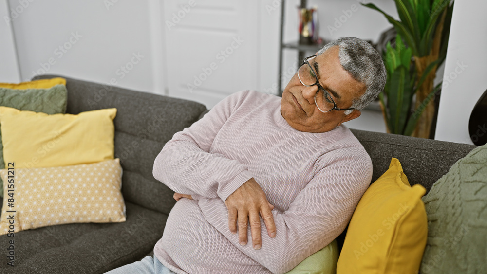A mature man relaxes on a couch at home, displaying comfort and tranquility in a modern living room setting. A mature man relaxes on a couch at home, displaying comfort and tranquility in a modern living room setting.