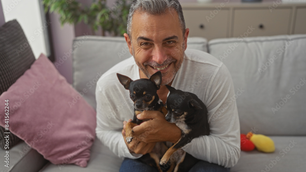 Obraz premium Middle-aged hispanic man holding two chihuahuas while sitting on a couch in a cozy living room.