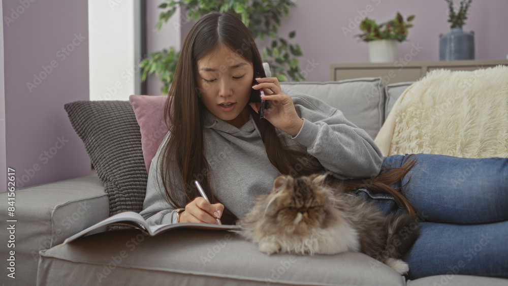 A young chinese woman lying on a couch in a living room, talking on the phone and writing in a notebook, with a fluffy cat resting beside her. A young chinese woman lying on a couch in a living room, talking on the phone and writing in a notebook, with a fluffy cat resting beside her.