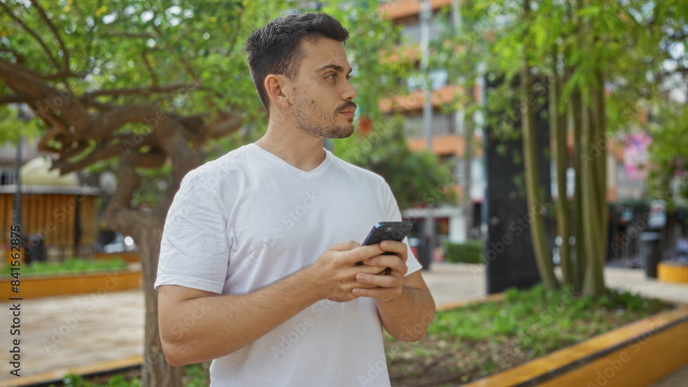 A handsome hispanic man with a beard standing in an urban park while holding a smartphone on a bright day outdoors. A handsome hispanic man with a beard standing in an urban park while holding a smartphone on a bright day outdoors.