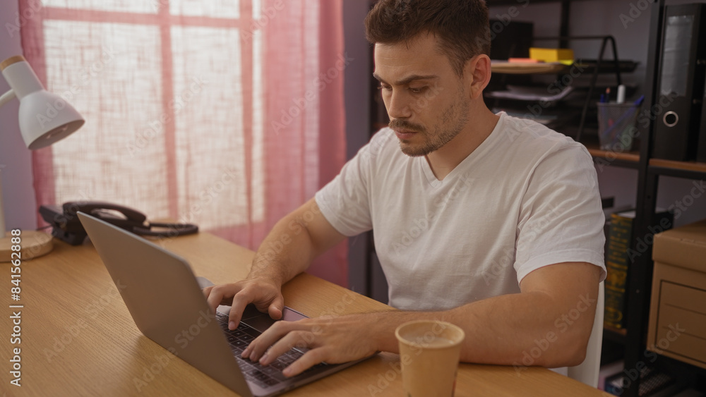 A young, handsome hispanic man working on a laptop in an office setting with a cup of coffee on the desk and bookshelves in the background. A young, handsome hispanic man working on a laptop in an office setting with a cup of coffee on the desk and bookshelves in the background.
