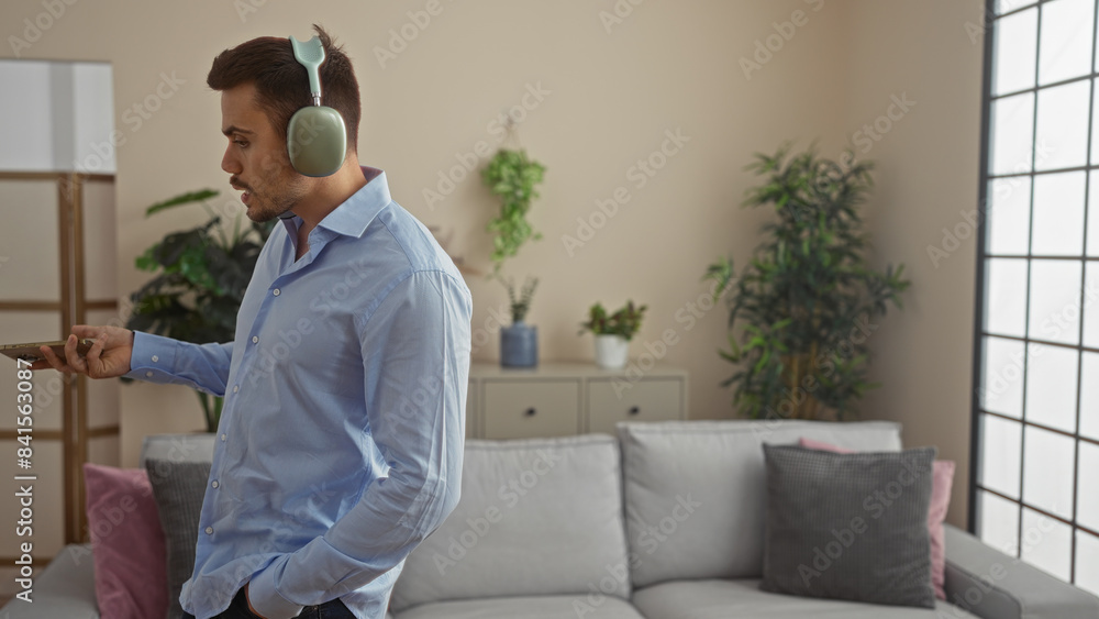 Young handsome hispanic man with a beard wearing headphones and holding a mobile phone stands in a modern living room apartment. Young handsome hispanic man with a beard wearing headphones and holding a mobile phone stands in a modern living room apartment.