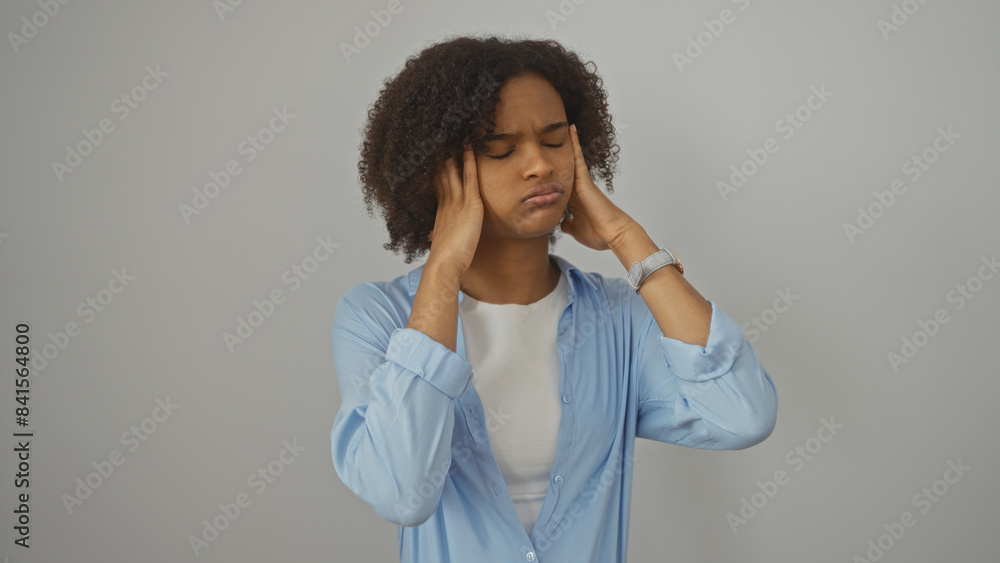 A young african american woman with curly hair stands against a white background feeling stressed and pressing her hands against her temples. A young african american woman with curly hair stands against a white background feeling stressed and pressing her hands against her temples.