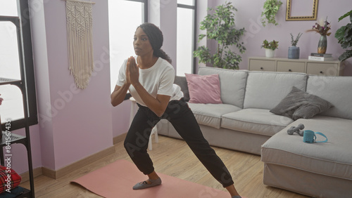 A young african american woman with curly hair performs a yoga pose in a cozy living room at home.
