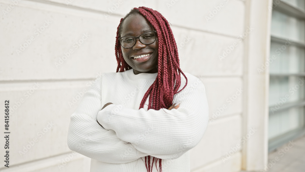 A smiling african woman with braids and glasses standing with arms crossed on an urban street. A smiling african woman with braids and glasses standing with arms crossed on an urban street.