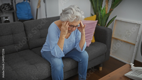 Elderly woman in glasses feeling stressed sitting on grey sofa indoors