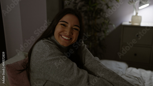 A smiling young hispanic woman sitting in a dimly lit indoor bedroom at night exuding warmth and comfort.