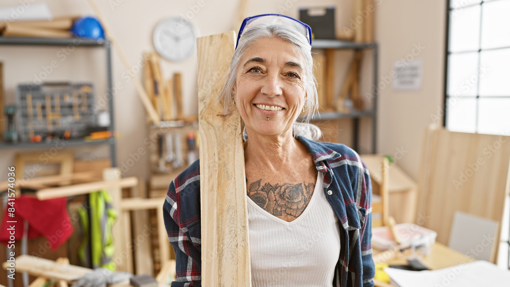 Smiling grey-haired middle age woman carpenter proudly holding a wood plank in her carpentry workshop Smiling grey-haired middle age woman carpenter proudly holding a wood plank in her carpentry workshop