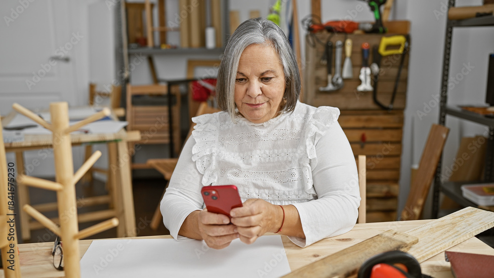Mature woman using smartphone in her woodworking studio filled with tools and wood Mature woman using smartphone in her woodworking studio filled with tools and wood