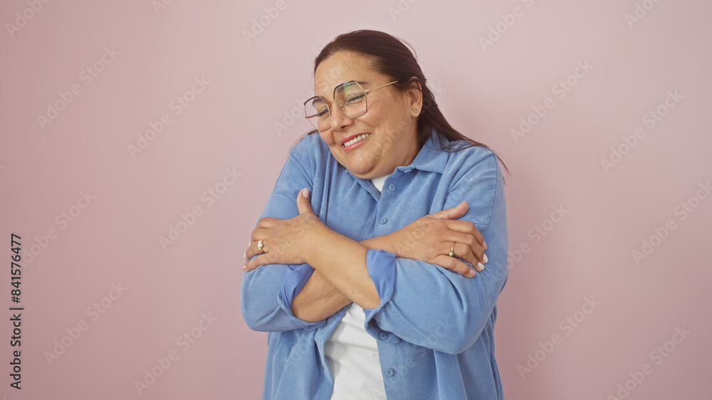 A smiling middle-aged hispanic woman hugs herself against a pink isolated background. A smiling middle-aged hispanic woman hugs herself against a pink isolated background.