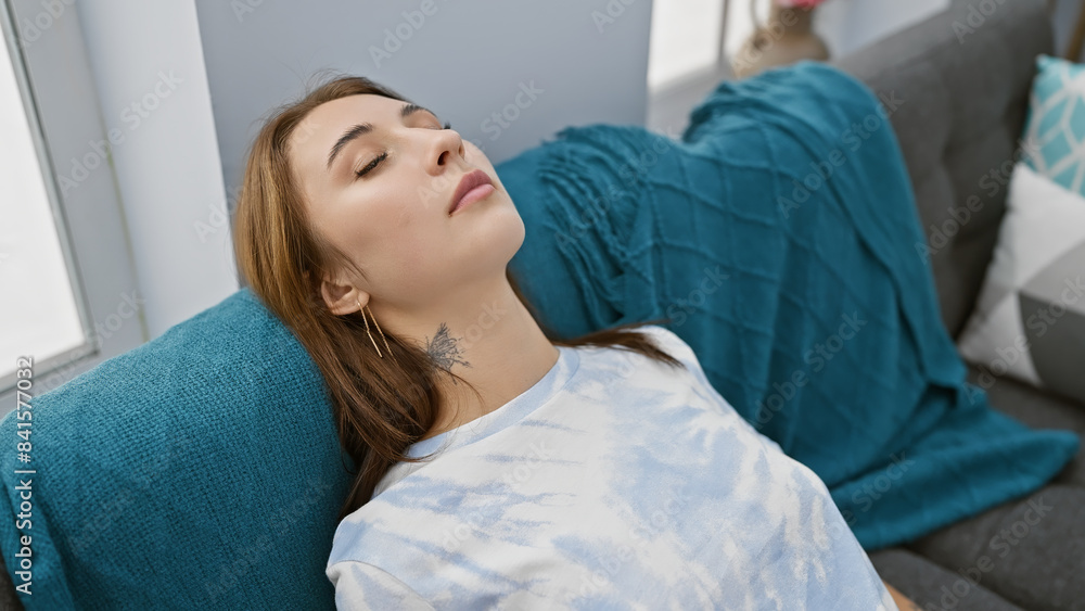 A serene young brunette woman relaxes on a sofa in a cozy living room, embodying casual comfort and modern home interior. A serene young brunette woman relaxes on a sofa in a cozy living room, embodying casual comfort and modern home interior.