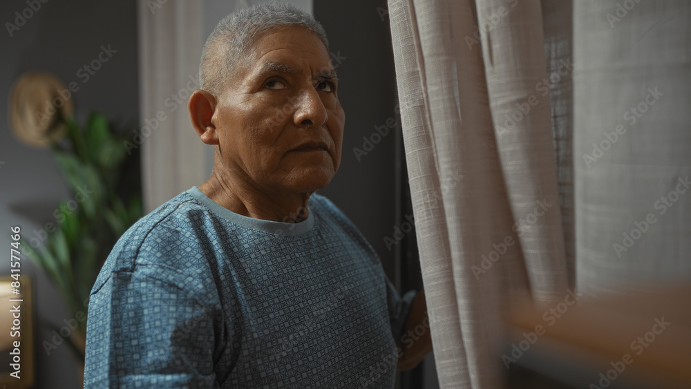 Hispanic senior man in a living room at home, standing near a window with grey hair and wearing a blue shirt, appearing thoughtful. Hispanic senior man in a living room at home, standing near a window with grey hair and wearing a blue shirt, appearing thoughtful.