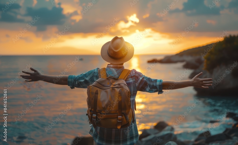Fototapeta premium Back view of a happy traveler man with a backpack and hat raising arms enjoying the sunset on the beach - standing with hands up looking sunrise - Self-care, wellness and healthy lifestyle concept