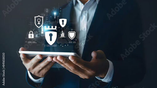 A businessman holds a tablet with shield and castle icons floating above it 