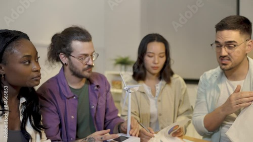A group of four young people from diverse backgrounds gather around a model wind turbine. They share their thoughts and ideas about sustainable energy sources. 