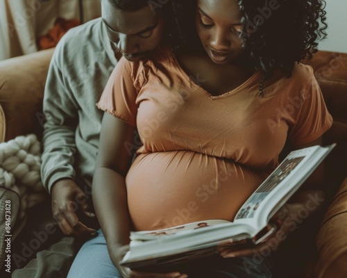 A pregnant woman sits on a couch with her partner, both looking down at a book in their laps. They are relaxed and cozy, enjoying each others company.